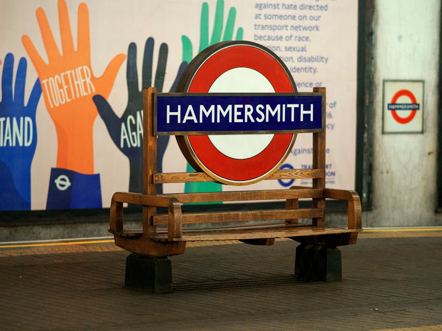 A wooden bench with a slatted seat and armrests, positioned inside a train station in front of a large circular red and white London Underground sign displaying the station name 'Hammersmith' in white