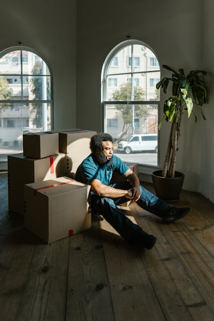 A man with curly black hair and a beard, dressed in a dark blue t-shirt and navy trousers, is inside a bright, unfurnished room with white walls and a large arched window behind him, natural light ill