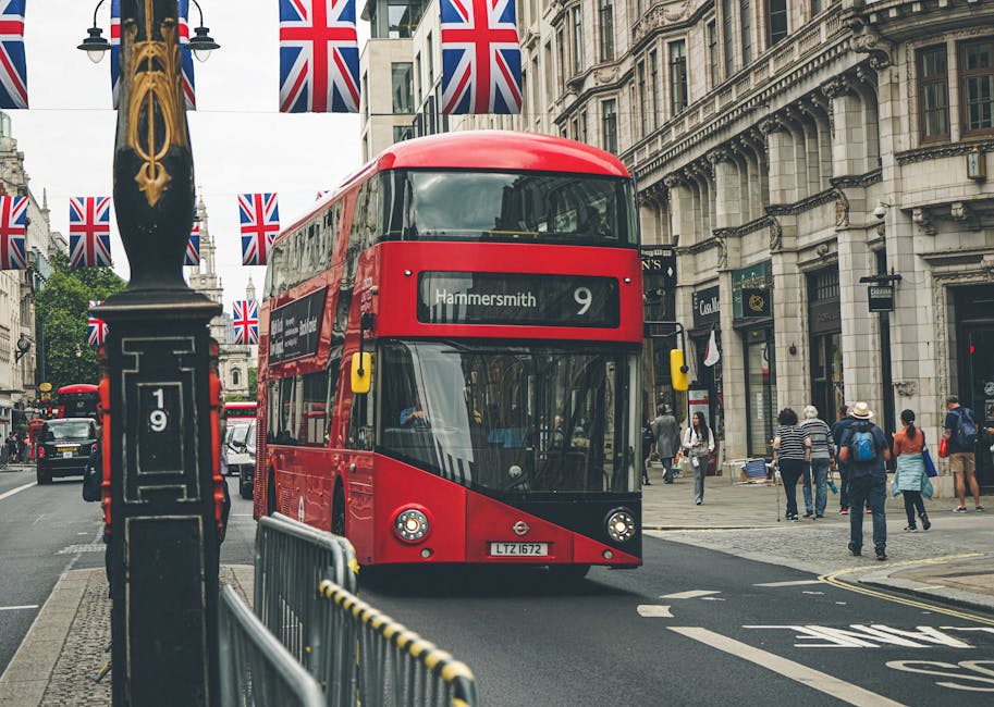 A red double-decker bus with route number 9 and destination 'Hammersmith' is driving along a busy city street in London, surrounded by pedestrians walking on the sidewalk and crossing the road. The st